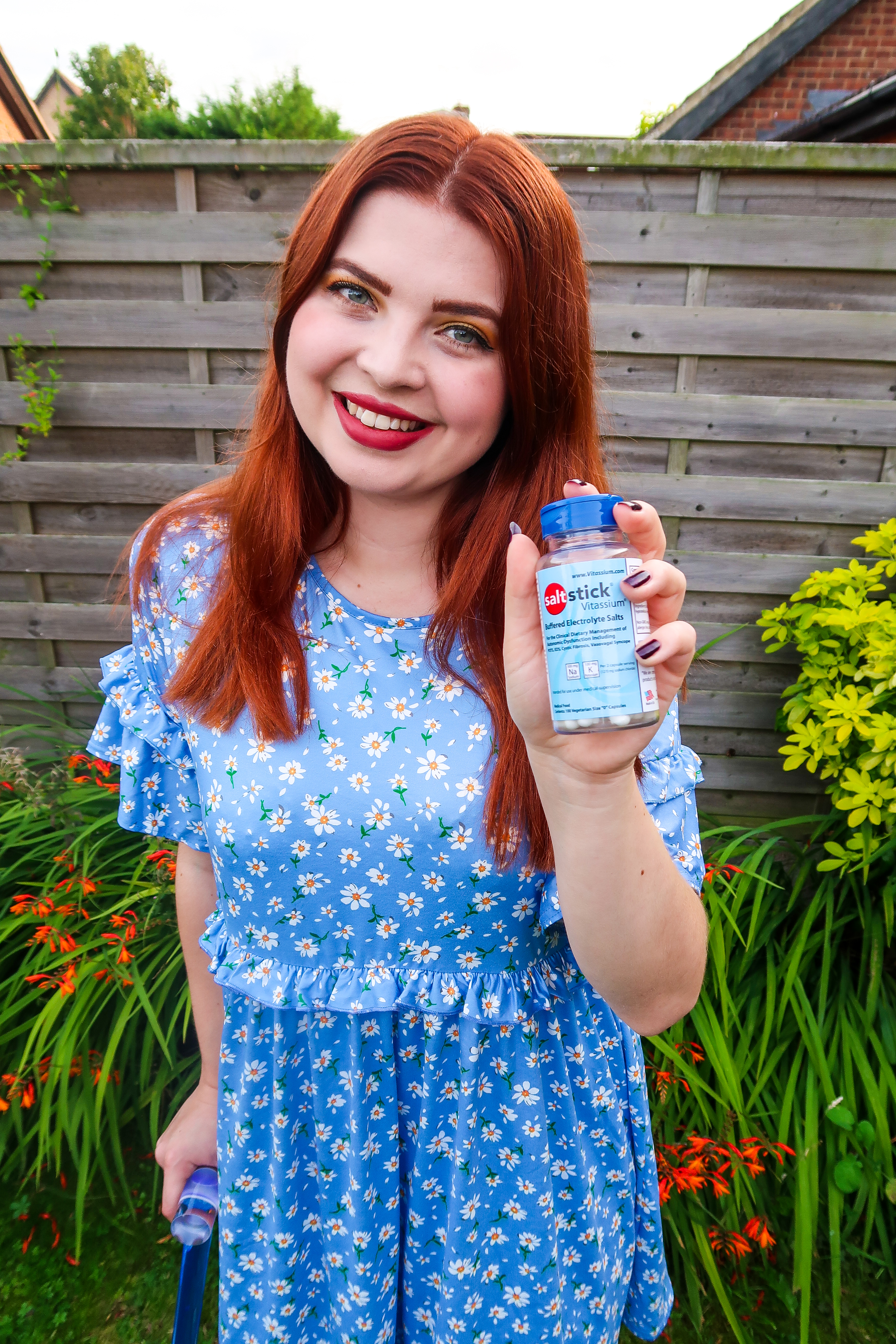 a midshot of Jenni a white woman with straight auburn hair is wearing a light blue dress with a small daisy pattern she is holding a bottle of vitassium a clear bottle with a dark blue top and pale blue label. she is using a blue walking stick and standing in a garden surrounded by plants.