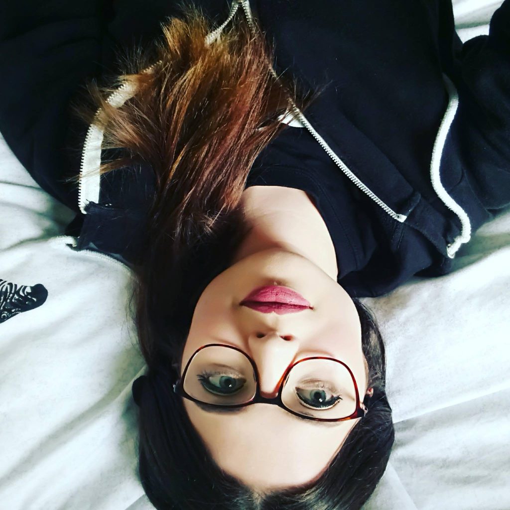 Jenni, laid upside down on a bed. She is wearing glasses and has pink makeup.