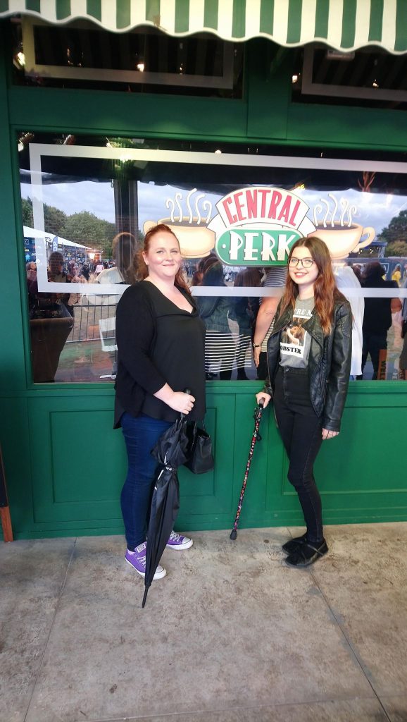 Jenni standing with her walking stick and another woman stood next to her with a purple walking stick. In the background is a glass window with the sign 'Central Park'. 