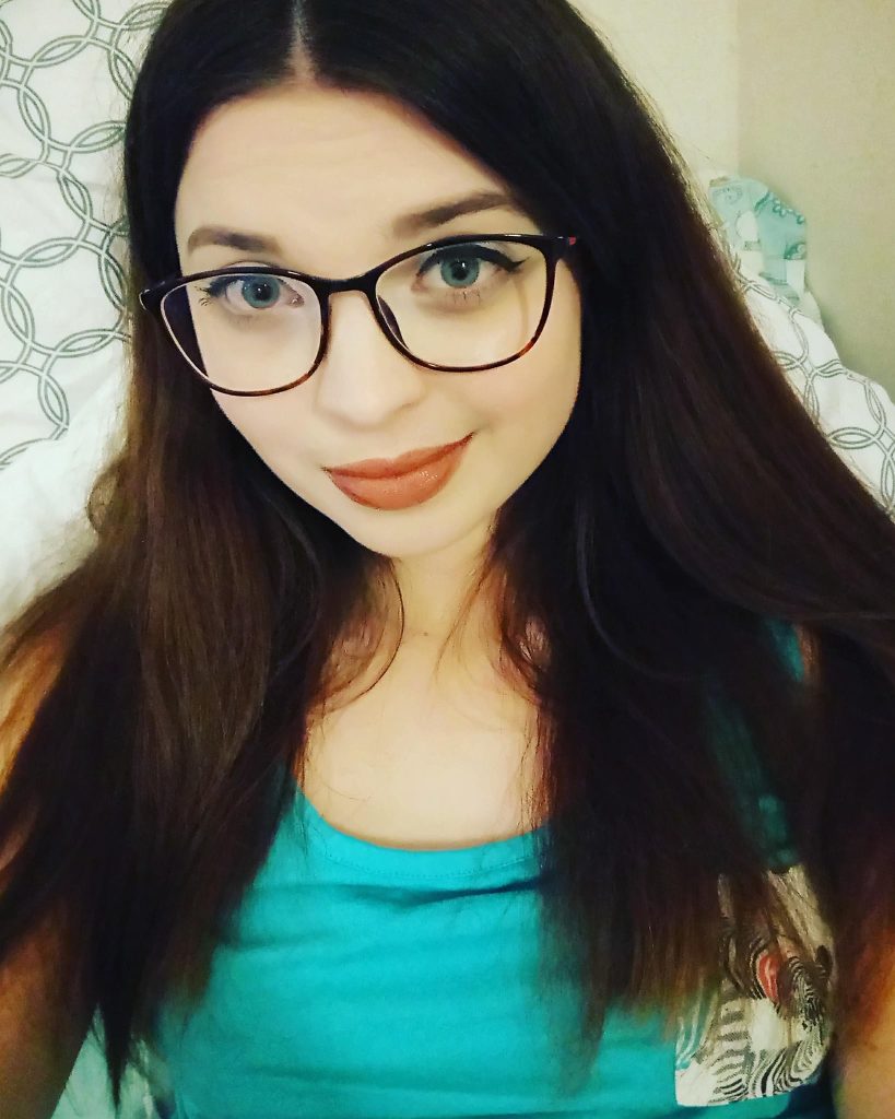 A selfie of Jenni sitting on a bed, wearing a blue top and glasses.