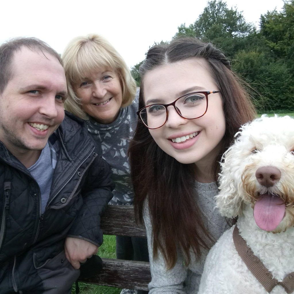 Jenni smiling at the camera, with a white, curly haired dog on her lap. Next to her is a man and woman who are also smiling.