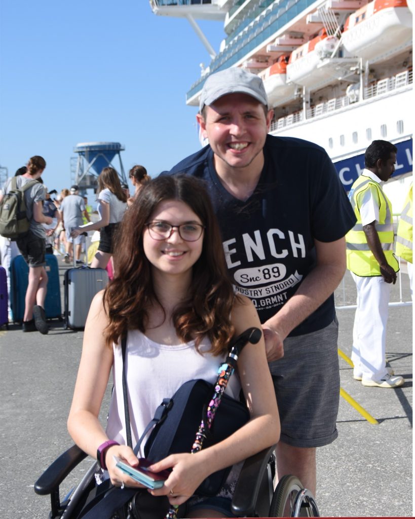 Jenni in a wheelchair in the sun, with a gentleman pushing her. 