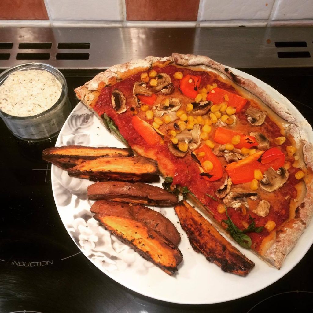 A plate with half a pizza which has toppings such as murshrooms and pepper, sitting next to some potato chips and dip.