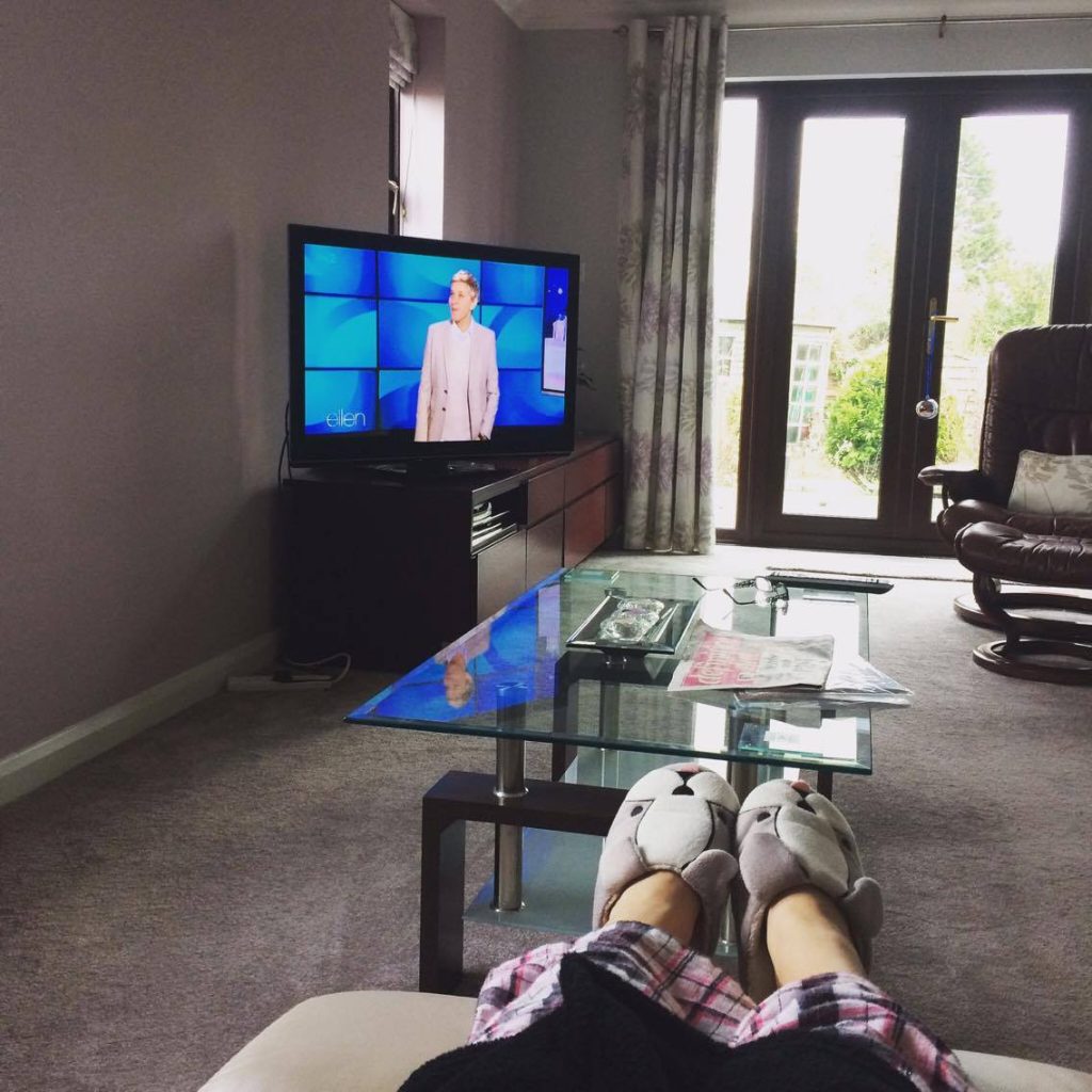 A POV photo of Jenni sat at home in her lounge, with the TV on. She is wearing her dog slippers.