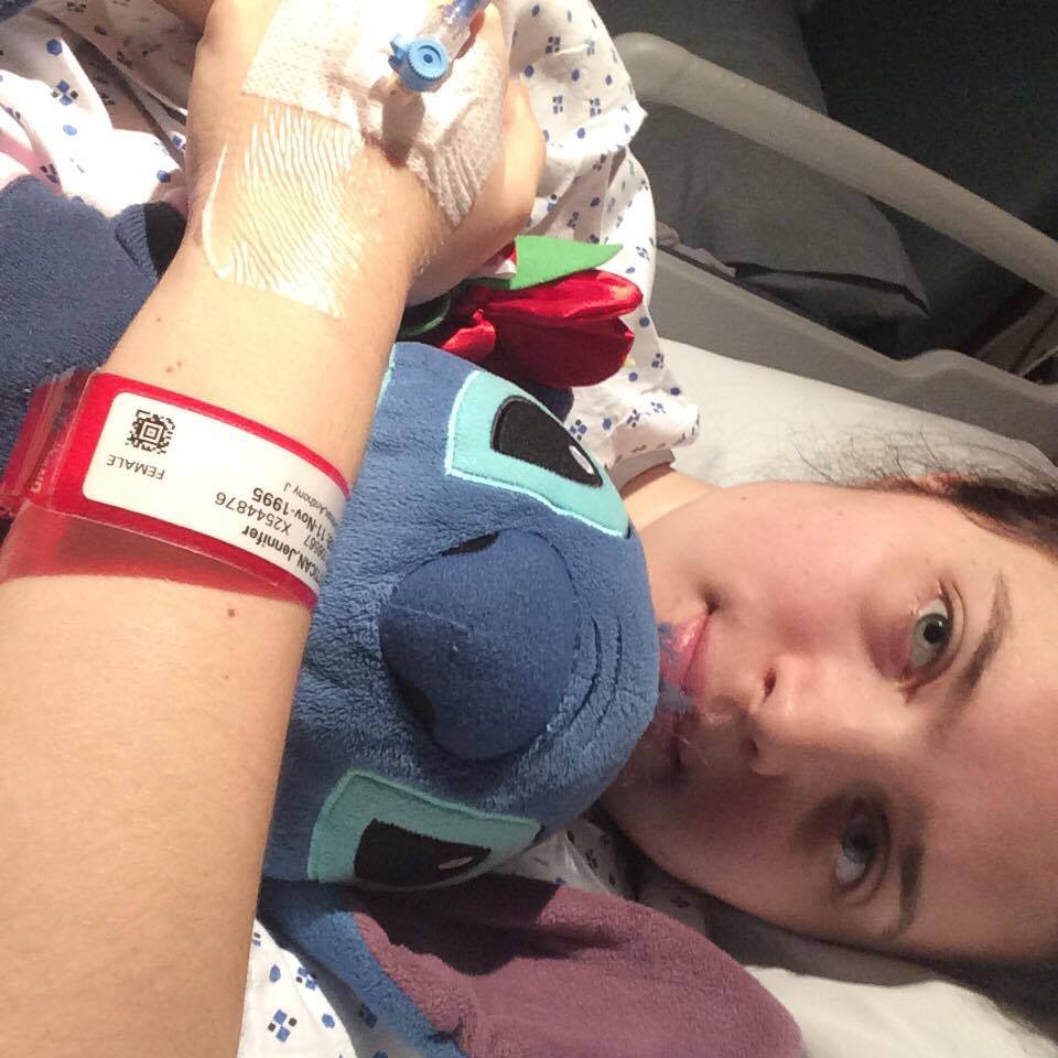 Jenni laid sidways in bed, looking blankly at the camera. A hospital band is visible, along with a Disney Stitch toy.