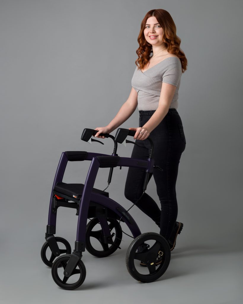 Jenni with wavy auburn hair smiles while standing beside a sleek purple rollator. She wears a fitted gray top and black jeans, posing against a plain gray background.