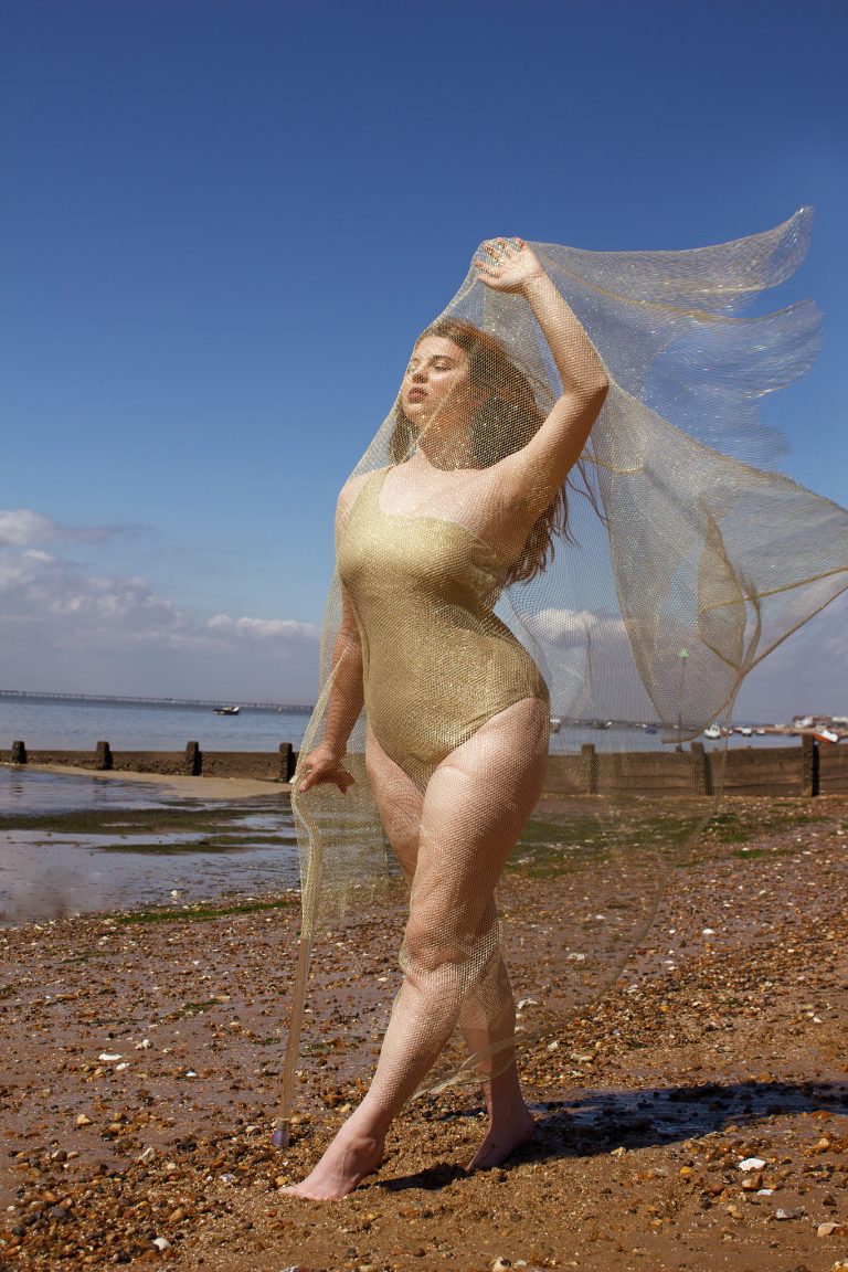 Jenni standing on a pebbled beach, wearing a gold swimsuit and draped in a sheer, shimmering fabric that catches the sunlight. She poses gracefully with one hand raised, her face serene, against a backdrop of a clear blue sky and calm sea.