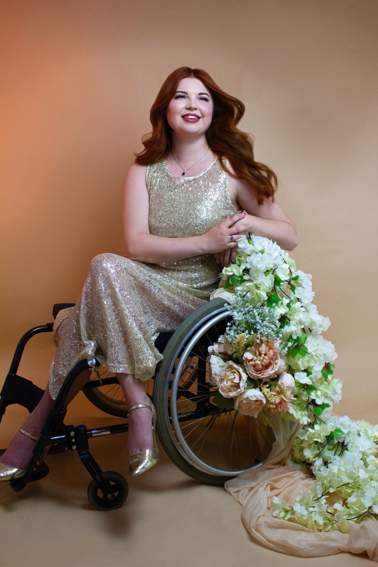 Jenni sat in her wheelchair, with flowers trailing off of it. She is wearing a gold sequin dress with heels.