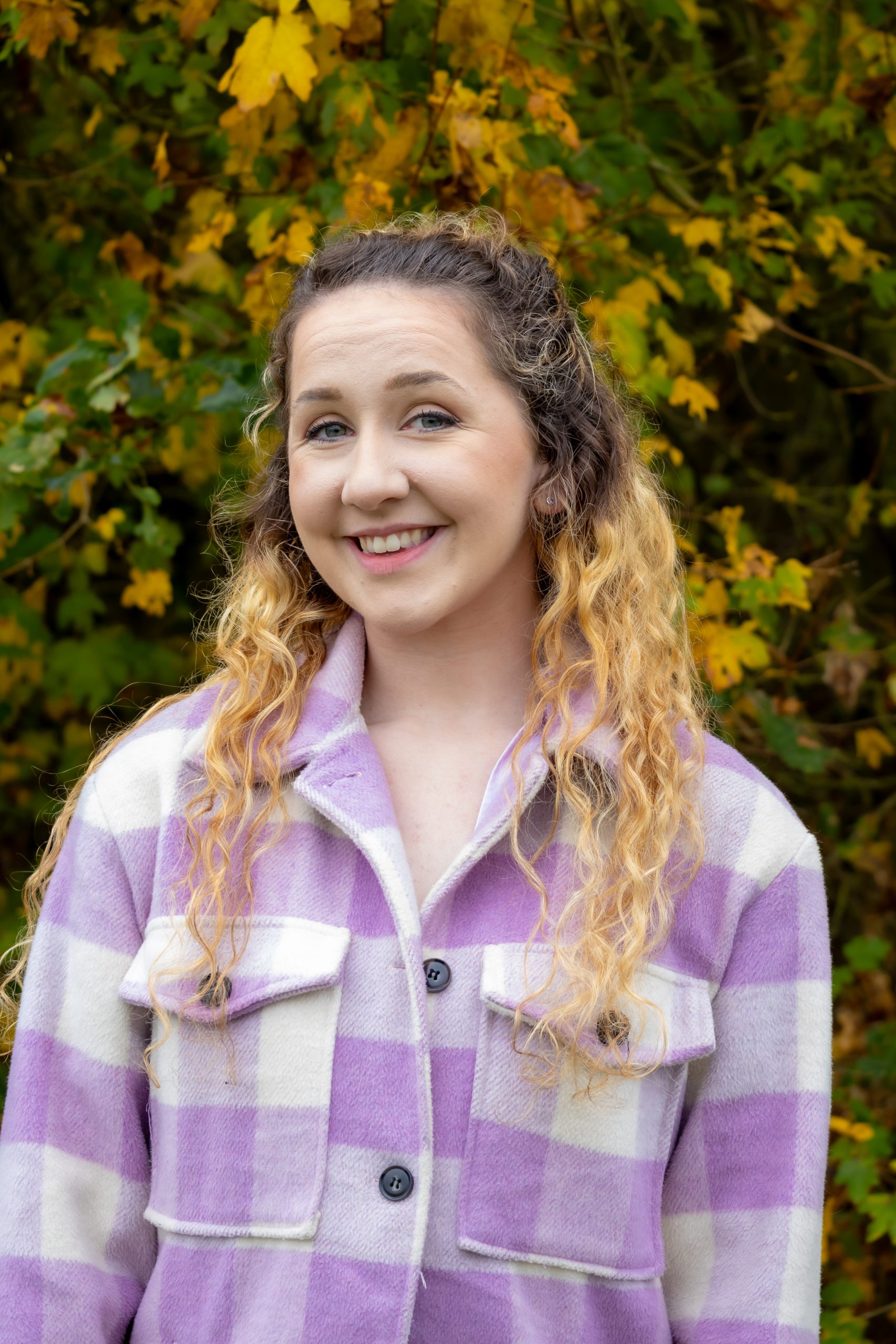 Beth smiles at the camera. She is wearing a purple plaid shacket and has long brown to blonde curly hair.