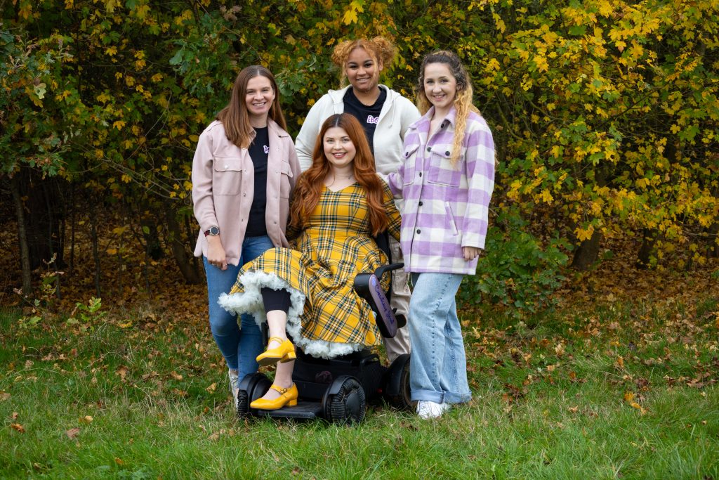 A group photo of the three team members, plus Jenni. Jennis wheelchair is sat in the middle of the three, with trees in the background.