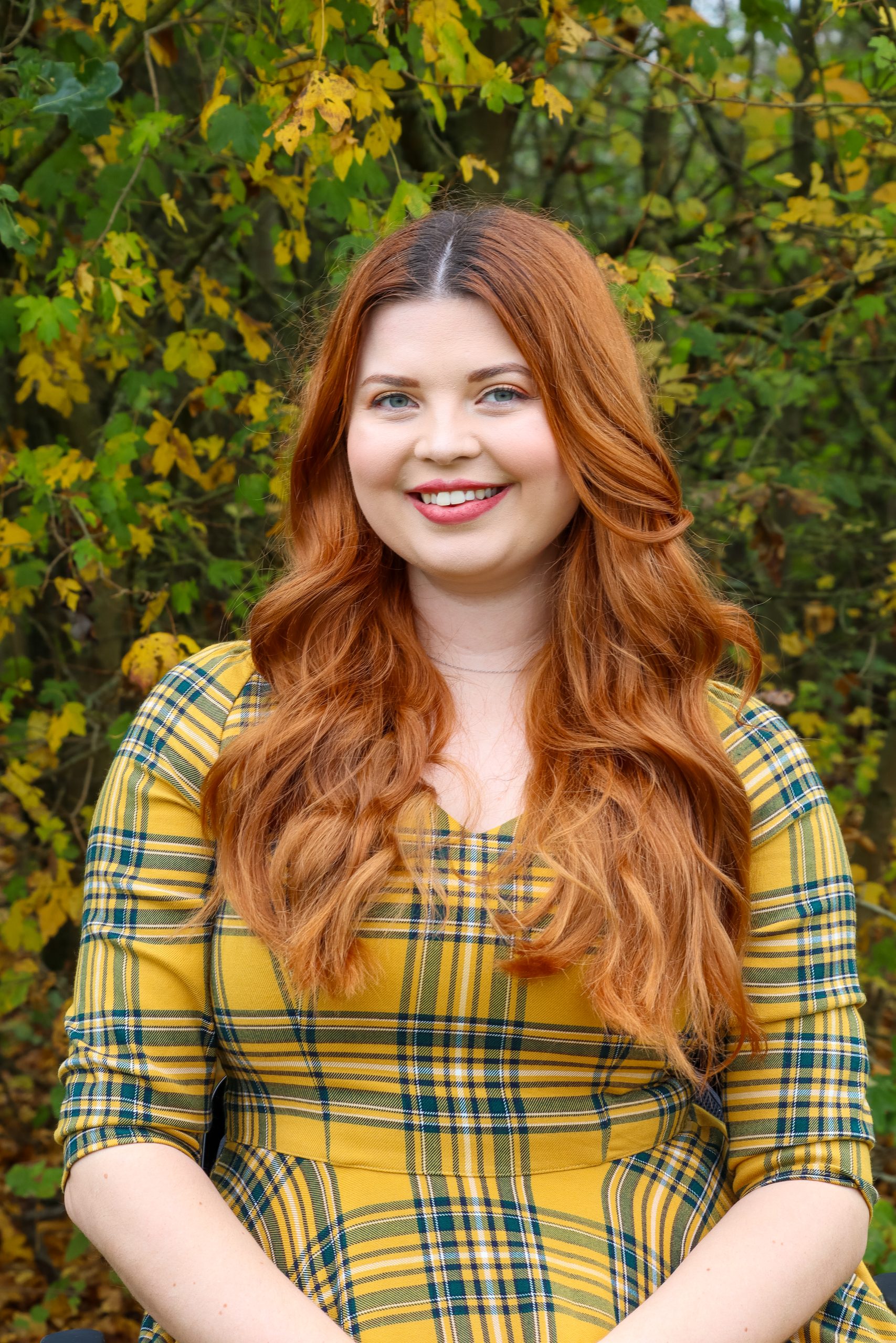 Jenni, a woman with long, auburn curled hair, smiling at the camera. She is wearing a yellow plaid dress, and has her arms on her lap.