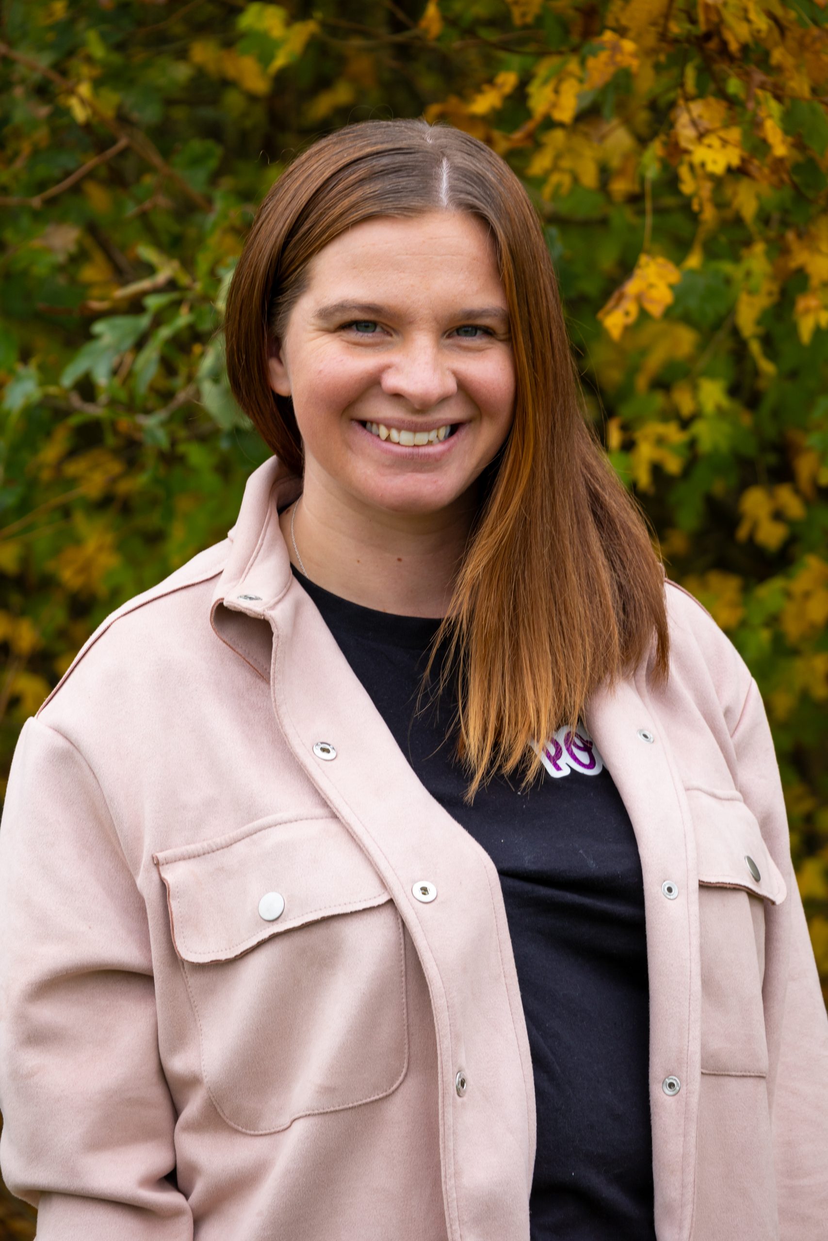 Poppy, a woman with long brown, straight hair smiles at the camera. She is wearing a black top and a pink shacket.