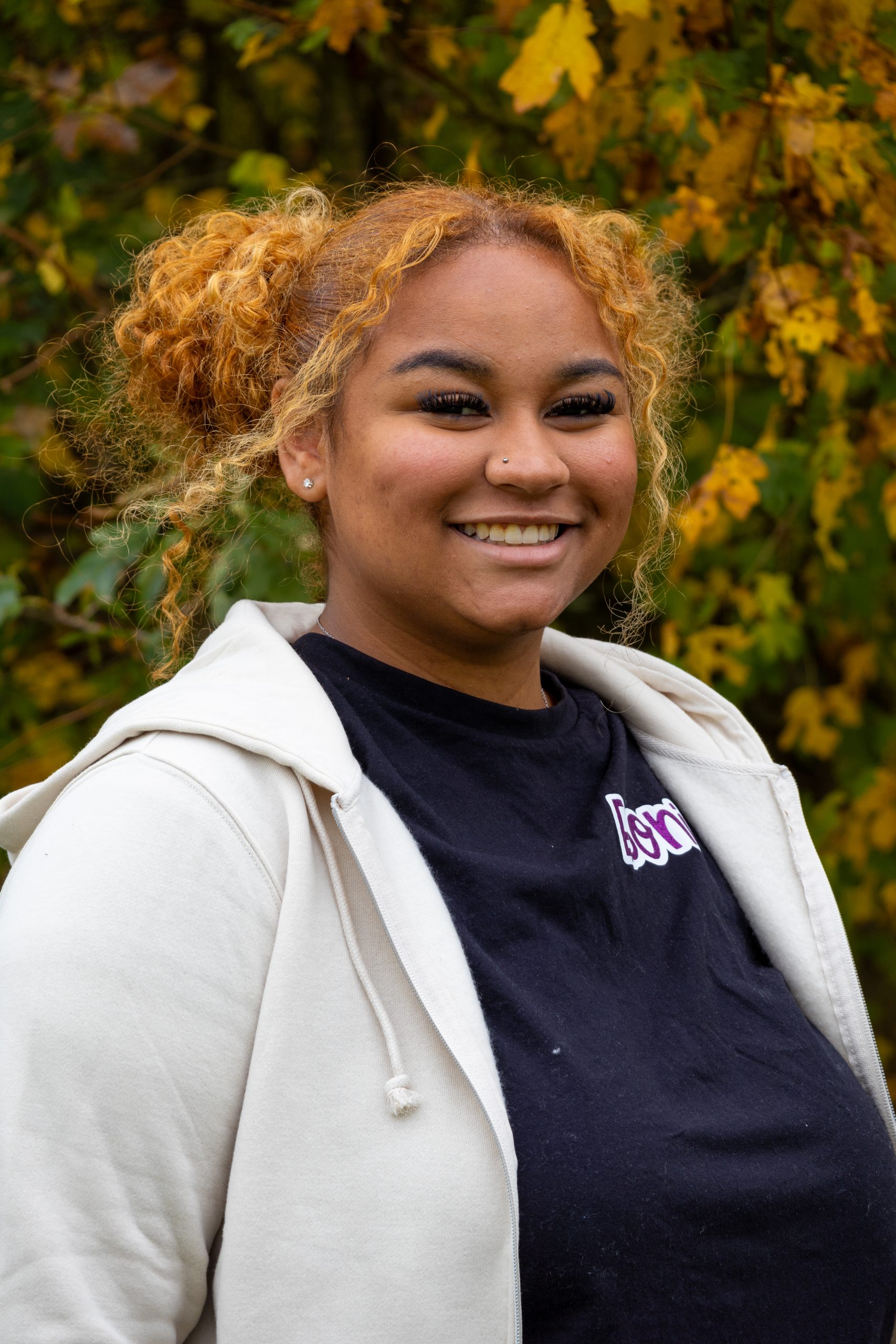 Eboni, a woman with blonde, curly hair pulled into space buns smiles at the camera, while wearing a black top and a grey hoodie.