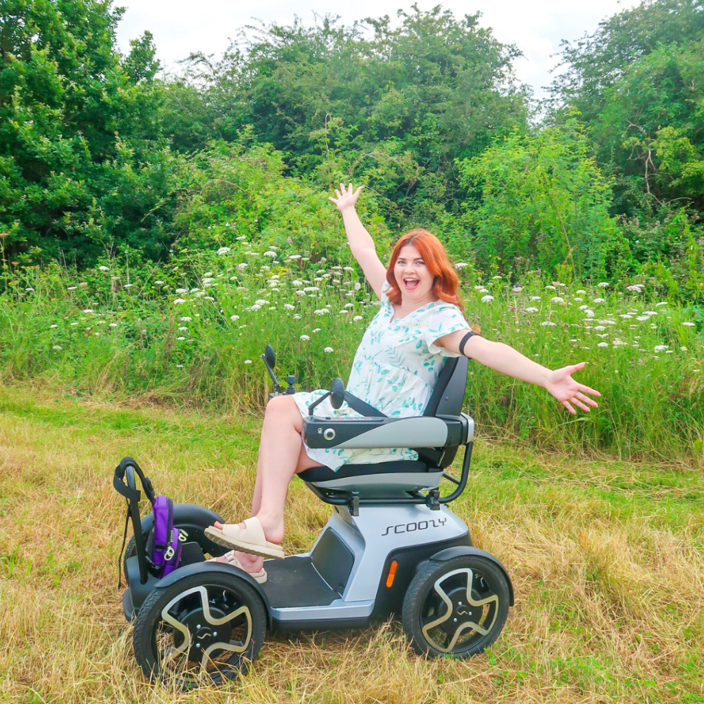 Jenni in her Scooty in a grassy field, with her arms out, smiling.