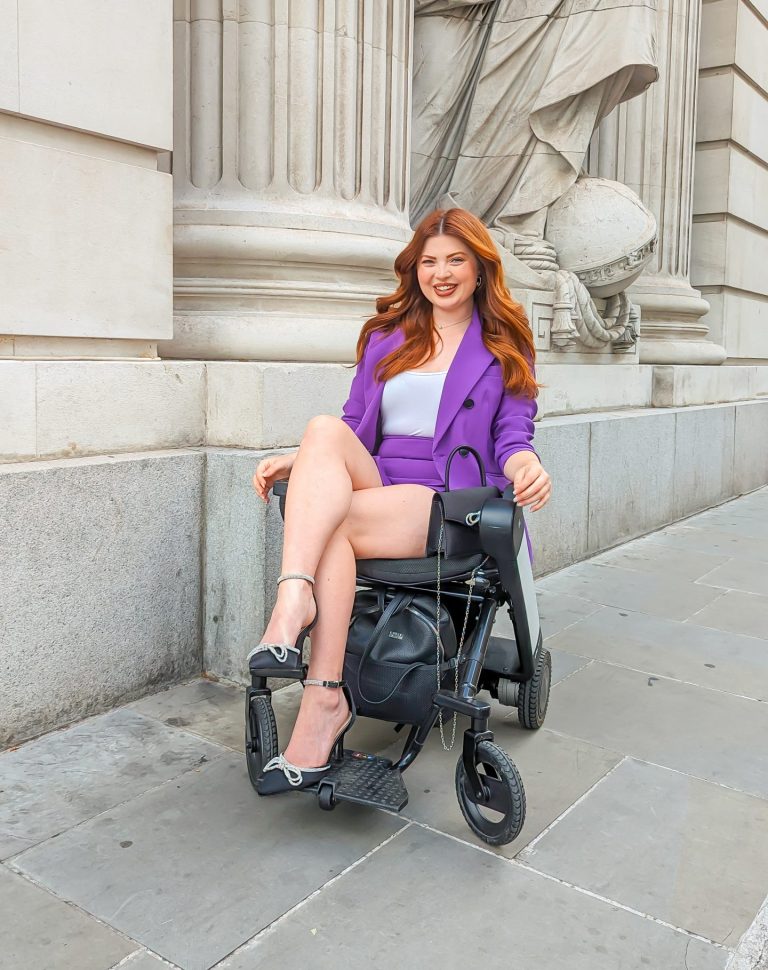 Jenni sitting in a wheelchair, wearing a purple suit with a grey top. Her long wavy auburn hair is falling around her shoulders. She is smiling.