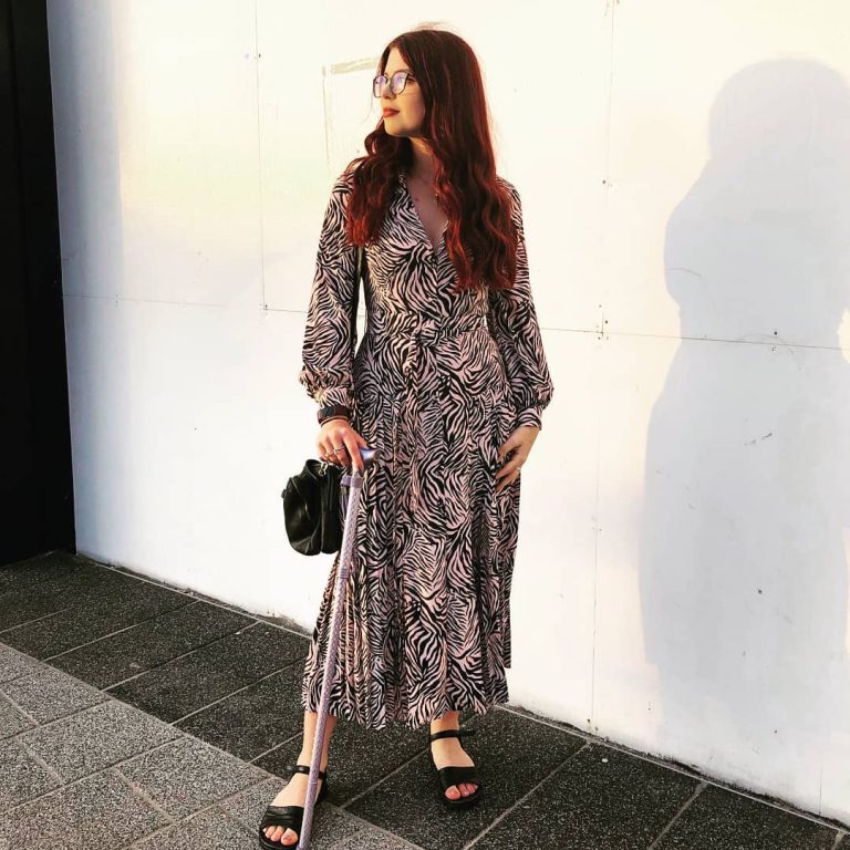 Jenni modelling a black and white zebra print dress next to a plain wall. The sun is casting a shadow behind her. She is leaning on her stick.