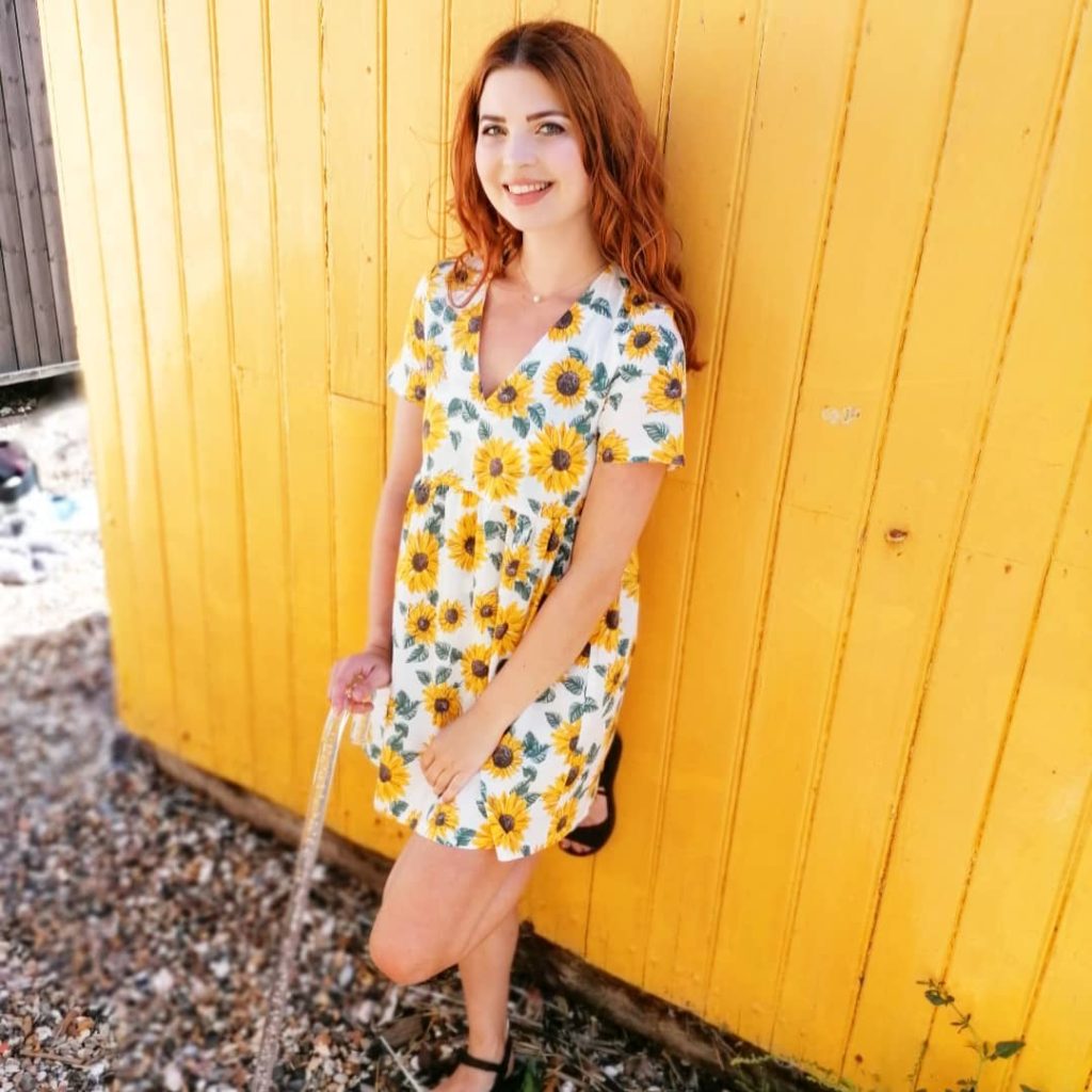 Raising mental health awareness, Jenni wearing a sunflower print dress, leant against a yellow wall.