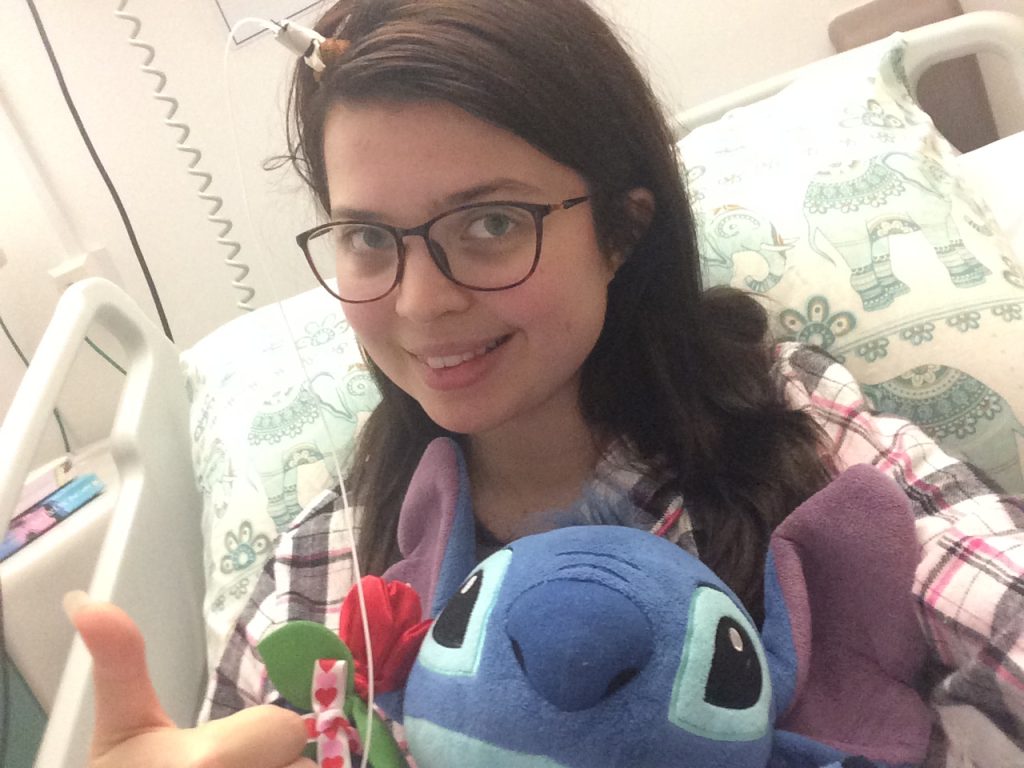 Jenni sitting in a hospital bed, smiling. She is holding a Disney Stitch toy and has visible machinery protruding from her head.