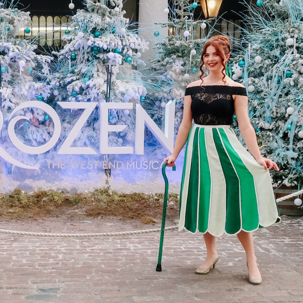 Jenni stood next to a size which reads Frozen The Musical. She is wearing a green striped skirt, with a black top and has her hair in a do. She is leaning on a green walking stick.