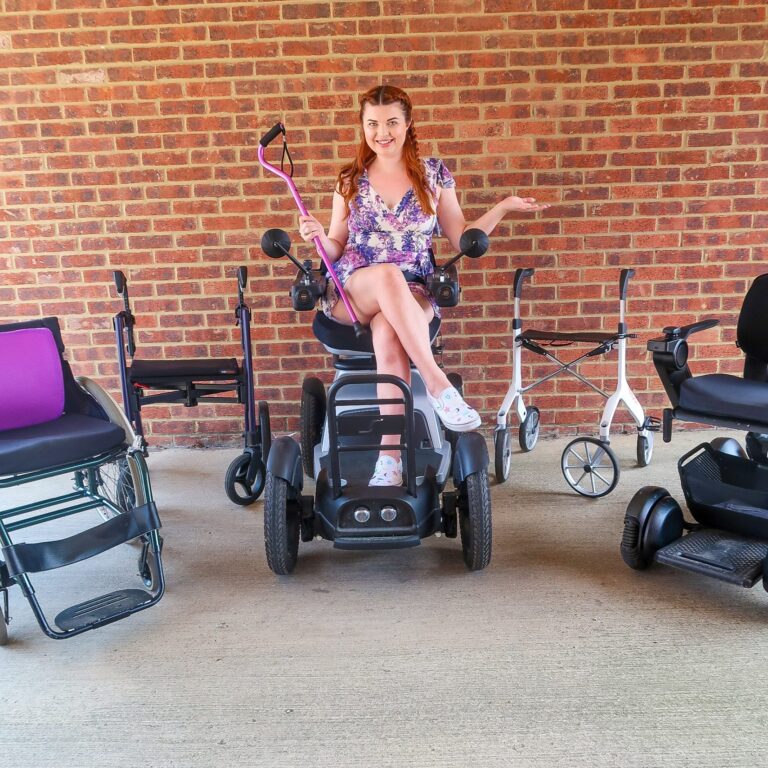 Jenni sitting in a wheelchair, holding a purple stick. Around her are various other mobility aids.