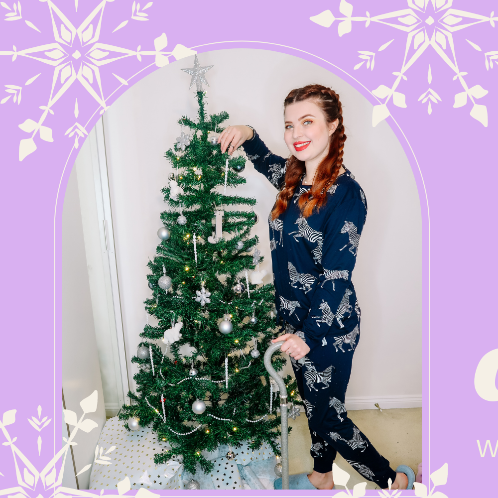 Jenni with long plaited red hair, wearing zebra pajamas, smiling at the camera. She is leaning against her spoonie holiday christmas tree, and her walking stick is visible.