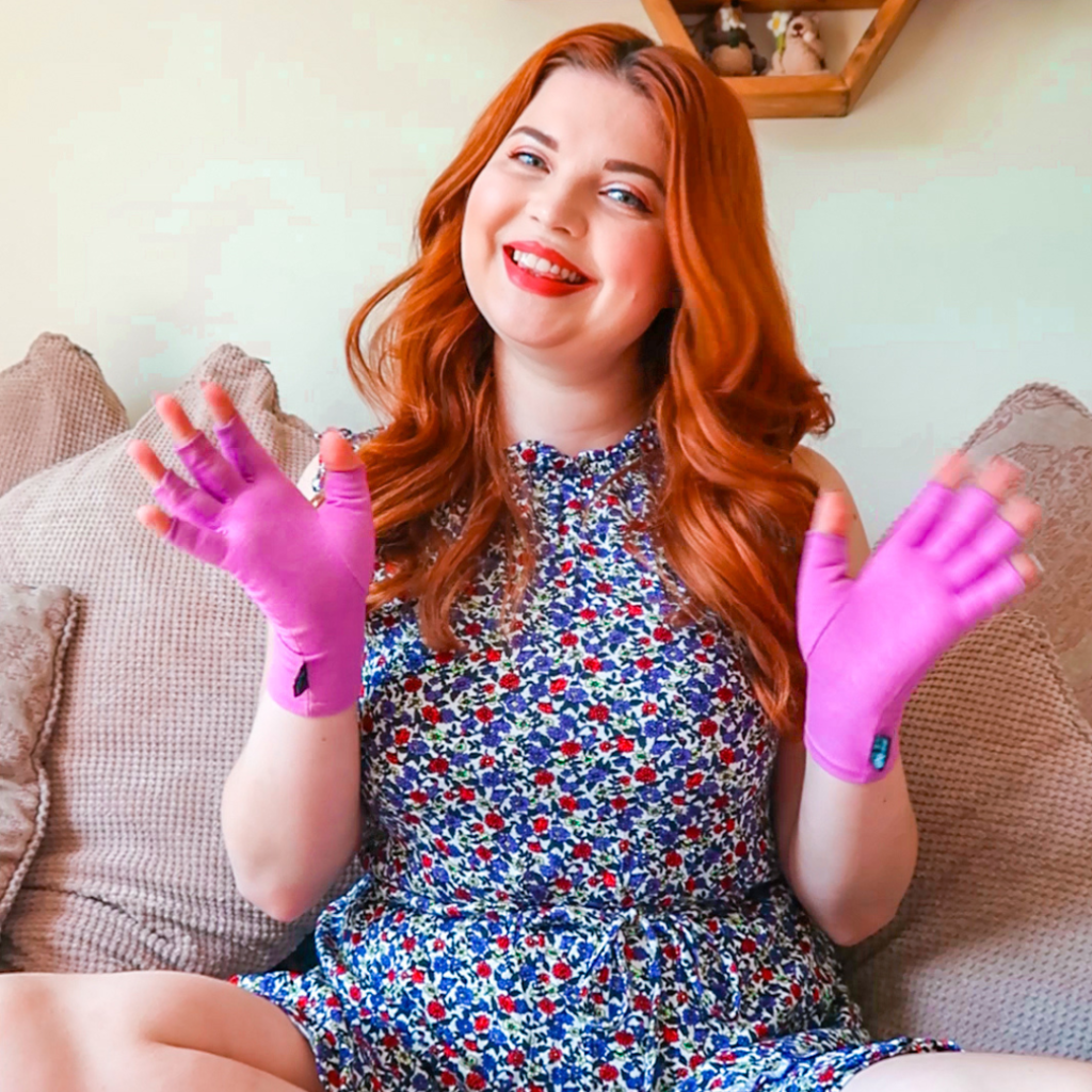 Jenni is sitting in a patterned dress on a grey sofa, wearing compression gloves - one of her favourite chronic-illness hacks.