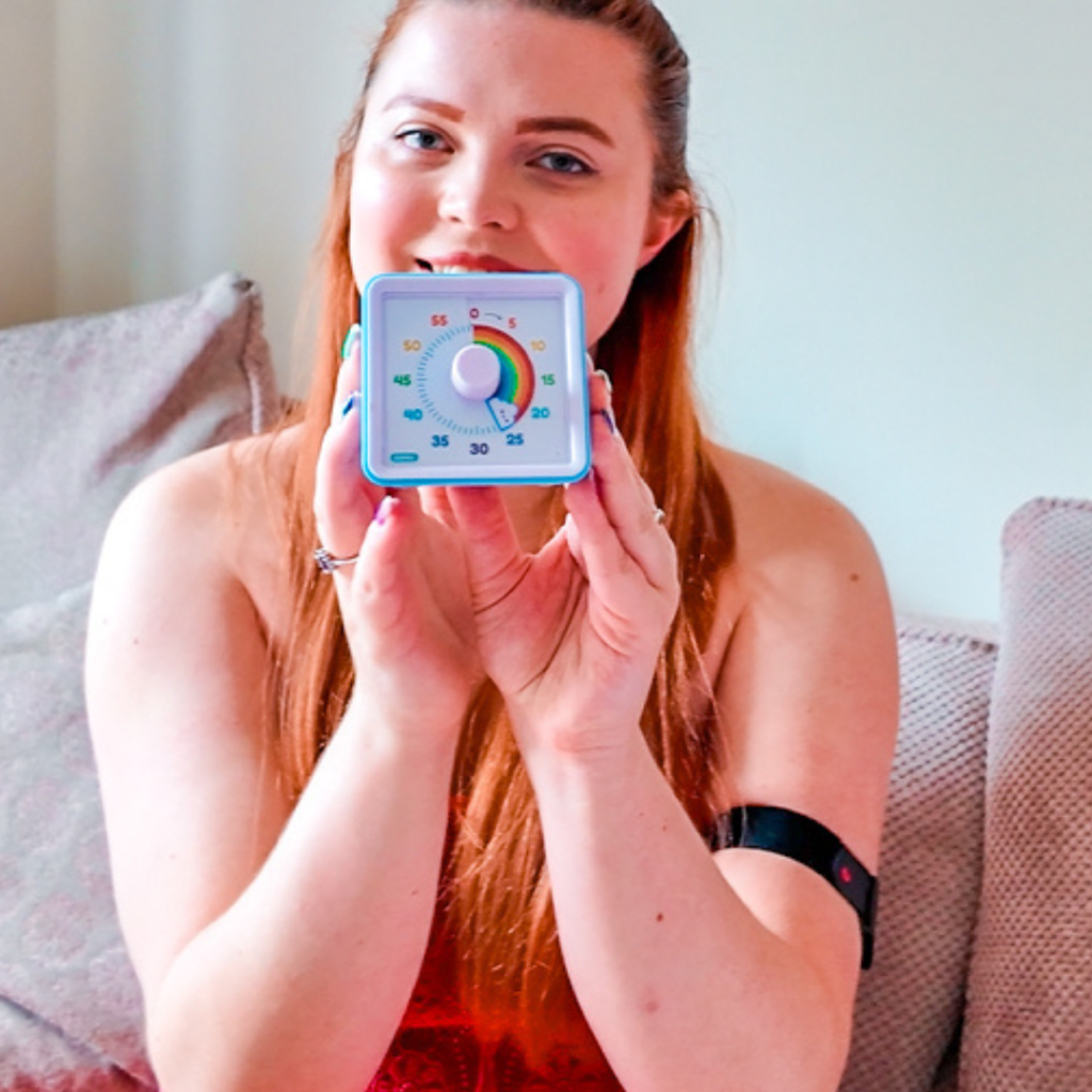 Jenni holding a timer up close to the camera. The timer has a rainbow to measure 20 minutes, after that, it has usual clock features. Jenni can be seen in the background smiling.