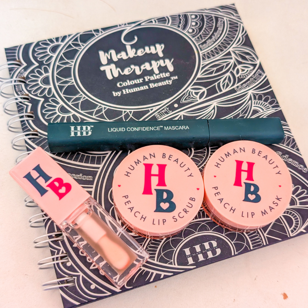 Human Beauty makeup products resting on a white desk. An eyeshadow palette, a lip oil, two bronzers and a mascara rest against each other.