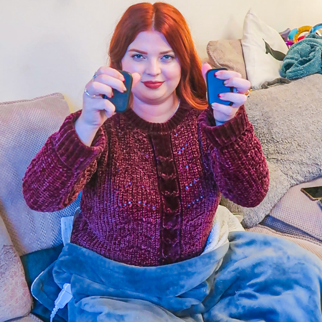 Jenni, sitting on a sofa, wearing a red jumper, holding up two hand heaters