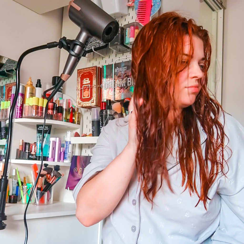 Jenni, sitting next to a hairdryer on a stand which is pointing at her hair, trying it. She is looking away from the camera.