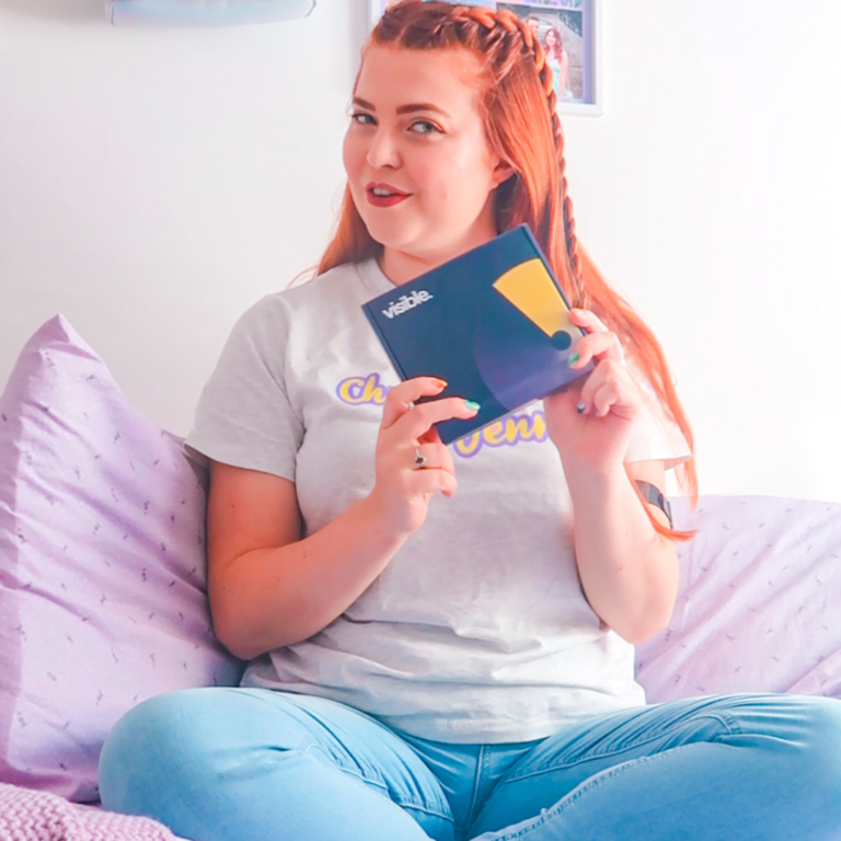 Jenni sat on a bed, holding a 'Visible' box. She is wearing a Chronically Jenni T-shirt and her hair is in part braids.