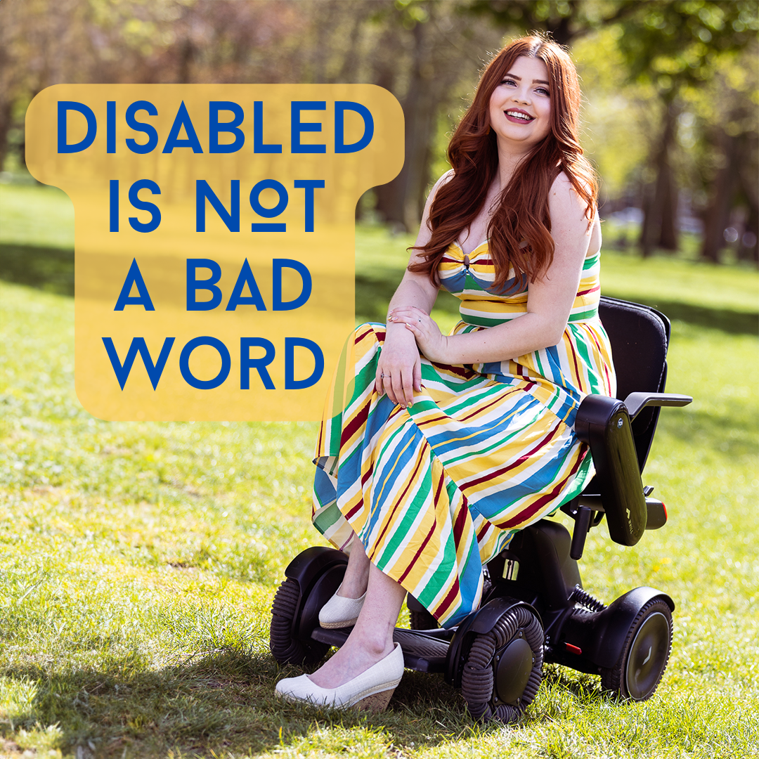 Jenni sat in her electric wheelchair in the park wearing a vertical striped red, yellow, blue, green and white dress and white wedges. The words Disabled is not a bad word are to one side of her