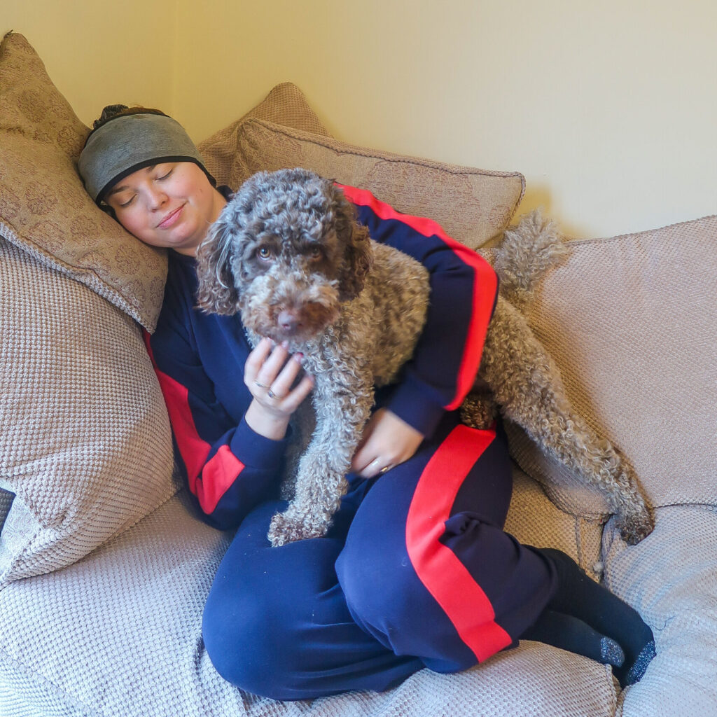 Jenni lying on the sofa wearing a headache headband with her eyes closed. Her labradoodle dog Coco is sat on her lap looking worried about her.