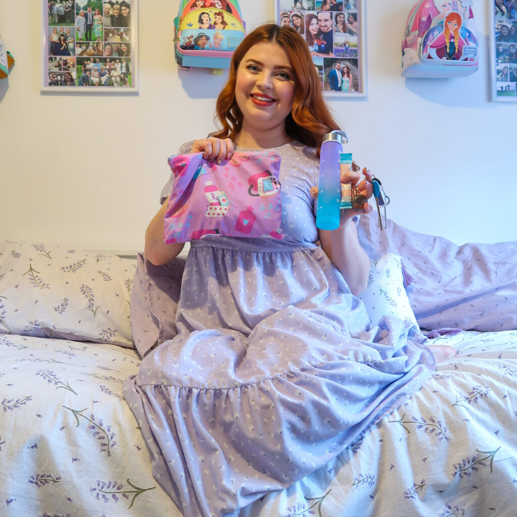 Jenni is sat on her bed holding her flare up bag in one hand and some items from inside it in the other including a small water bottle, a sachet of electrolytes, her loop earplugs and a radar key