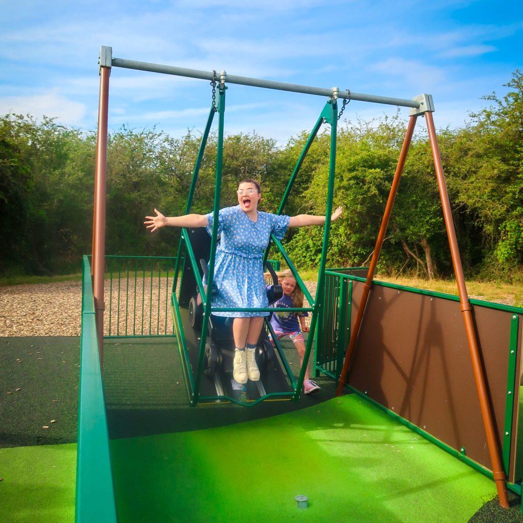 Jenni a disabled woman wearing a blue dress is sat in her electric wheelchair on an adapted swing at the park with her arms outstretched in delight. She is being push by a blonde 8 year old