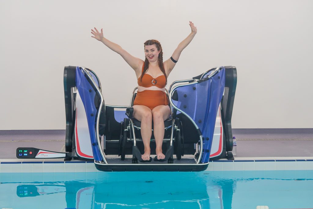 Jenni sat in a pool wheelchair in a swimming pool lift called a pool pod which is being lowered into the water of the swimming pool. She is wearing a rust coloured bikini and has her arms in the air in delight