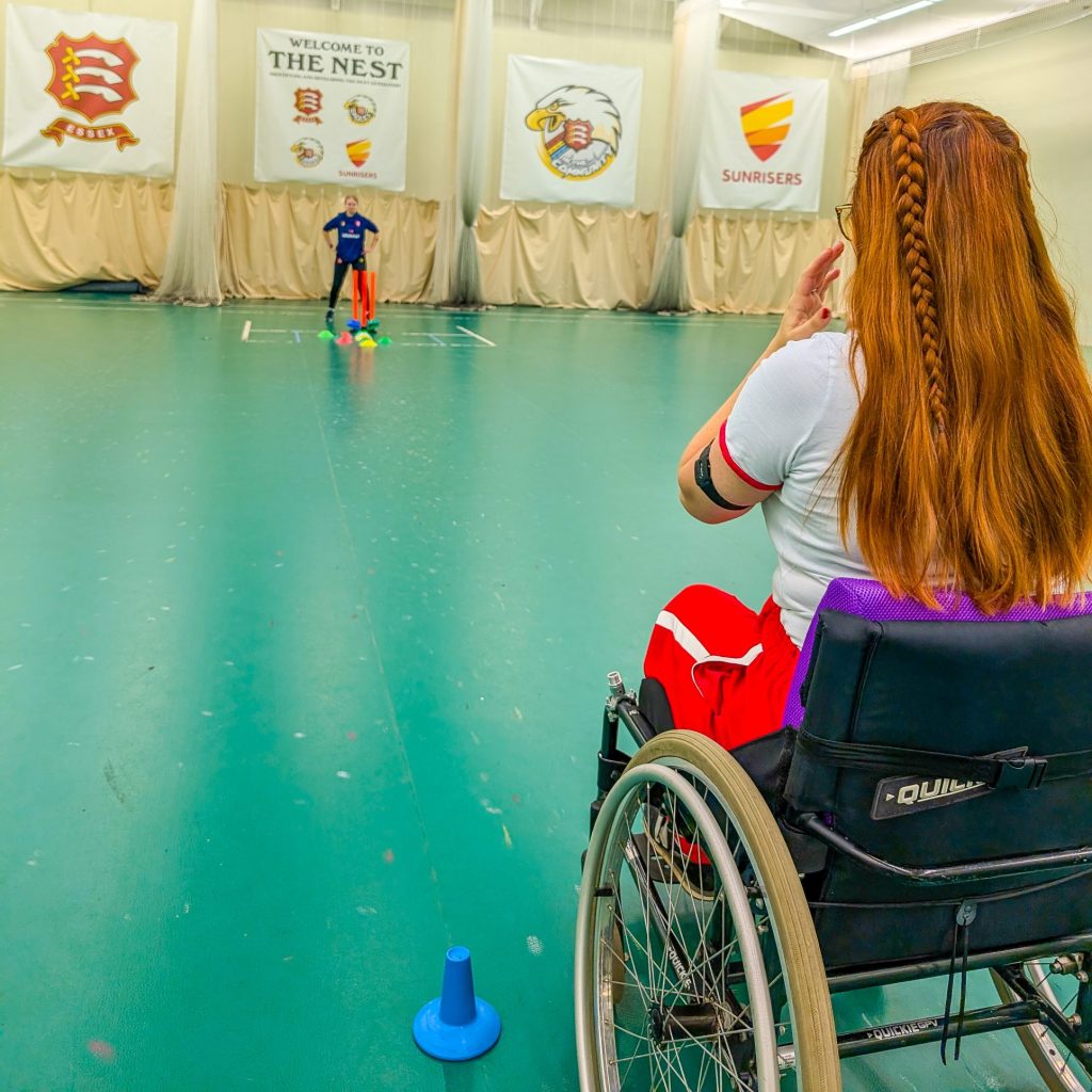 a shot of Jenni from behind sat in her manual wheelchair about to bowl a cricket ball at some wickets at an indoor cricket ground