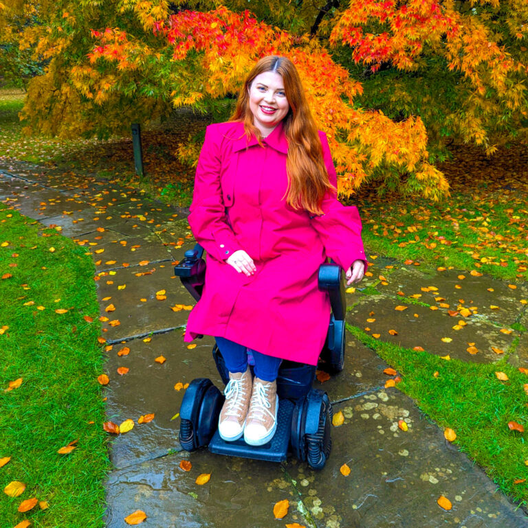 Jenni sat in an autumnal garden in her electric wheelchair wearing a bright pink adaptive coat