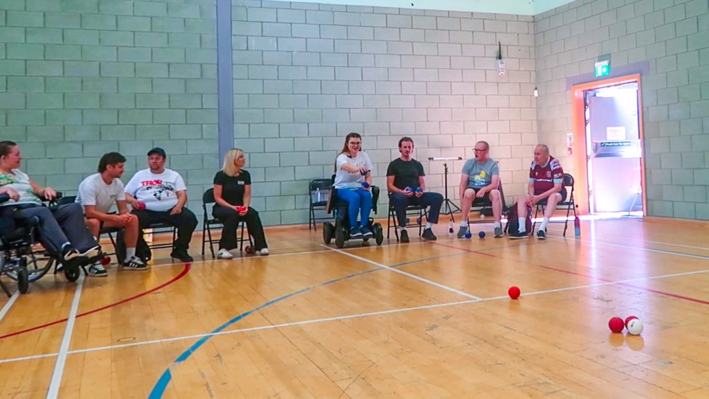 an photo of Jenni in a sports hall sat in her electric wheelchair with a group of other disabled and non disabled people all sat down playing boccia. Jenni is holding a blue boccia ball ready to throw towards the white jack which is surrounded by red team balls.