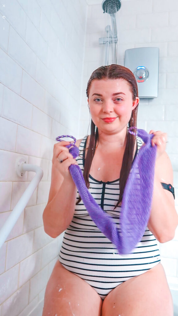 Jenni sitting in the shower wearing a swimming costume, holding a purple african exfoliating net