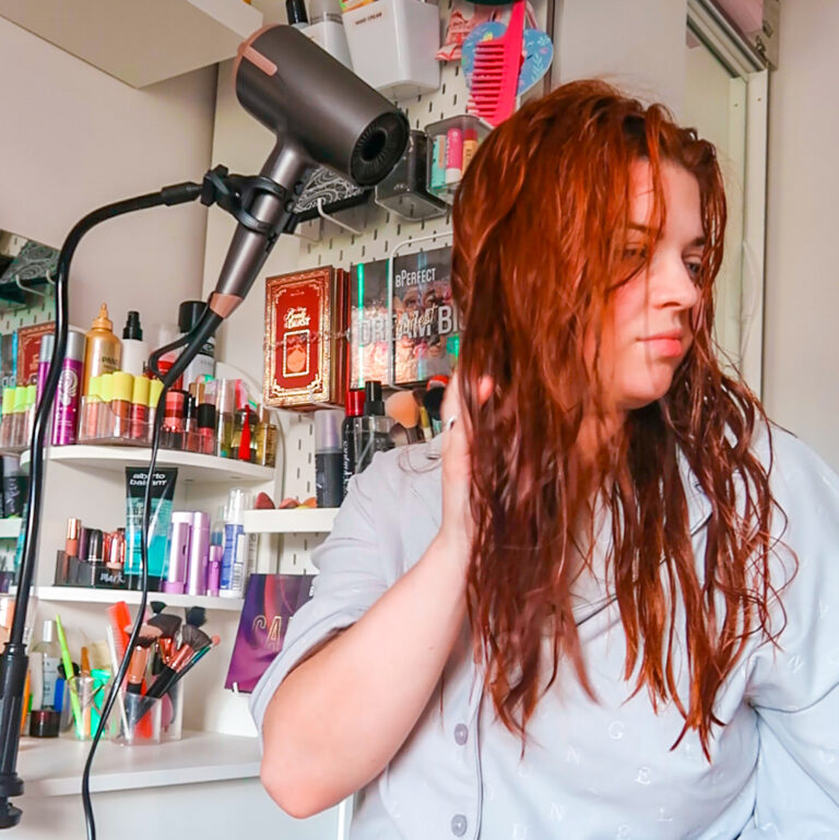 Jenni is drying her hair demonstrating her Disability Hacks. A black gadget is clipped to her dressing table and holding a grey hairdryer up on the other end drying Jenni's hair