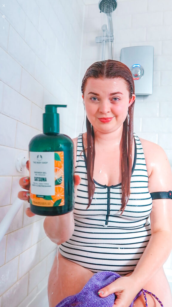 Jenni sitting in the shower wearing swimming costume holding a bottle of shampoo with an easy-pump lid.