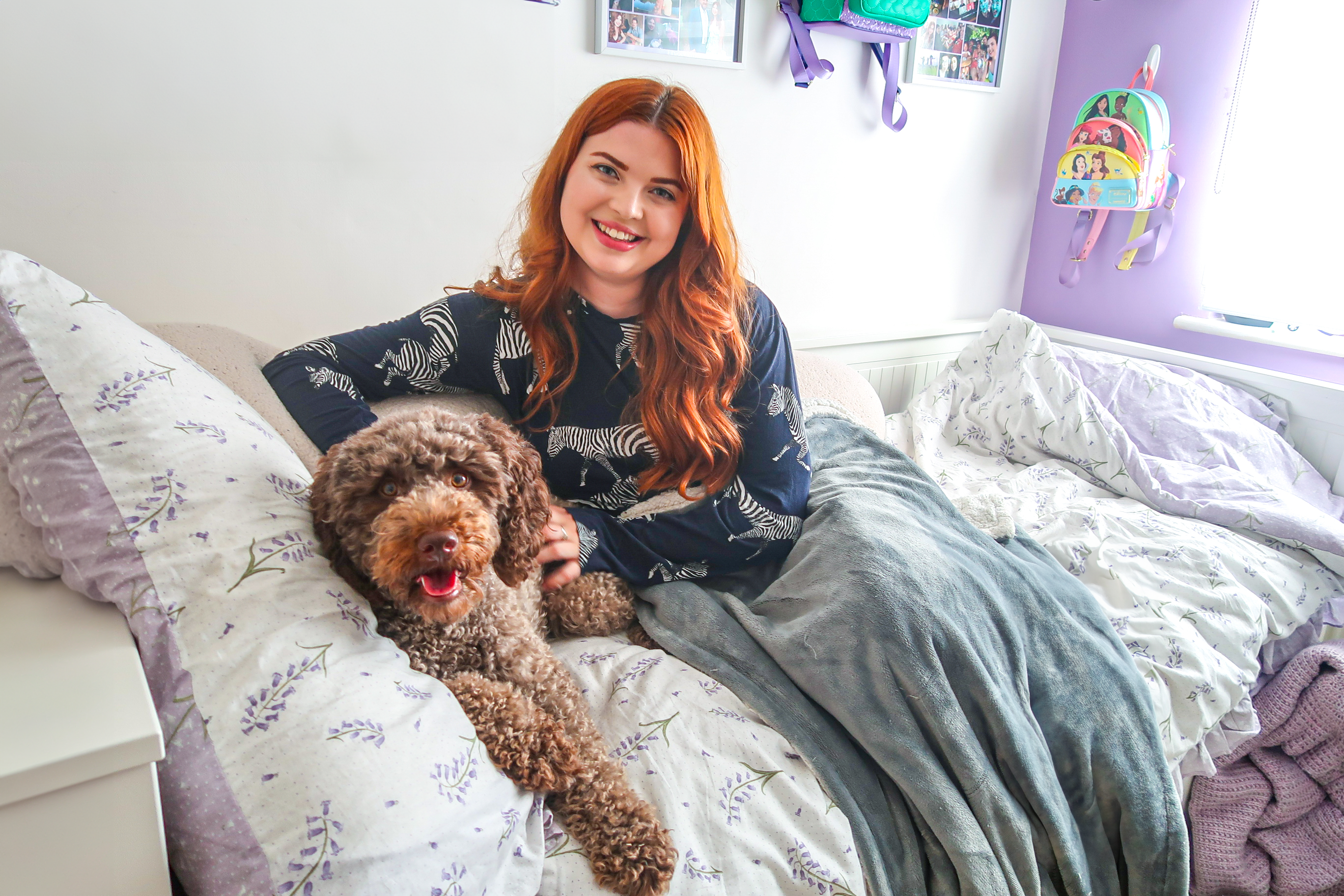 Jenni sitting on her bed wearing zebra pjs. She has her arm on her brown haired dog. She is smiling and looking at the camera.