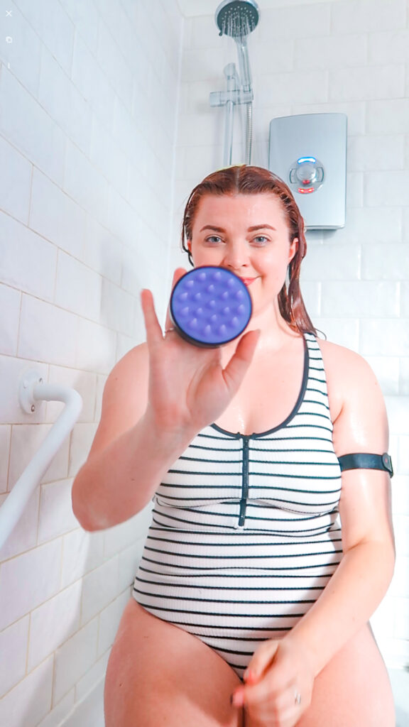 Jenni, sitting in the shower wearing a swimming costume, holding her silicone shampoo brush.