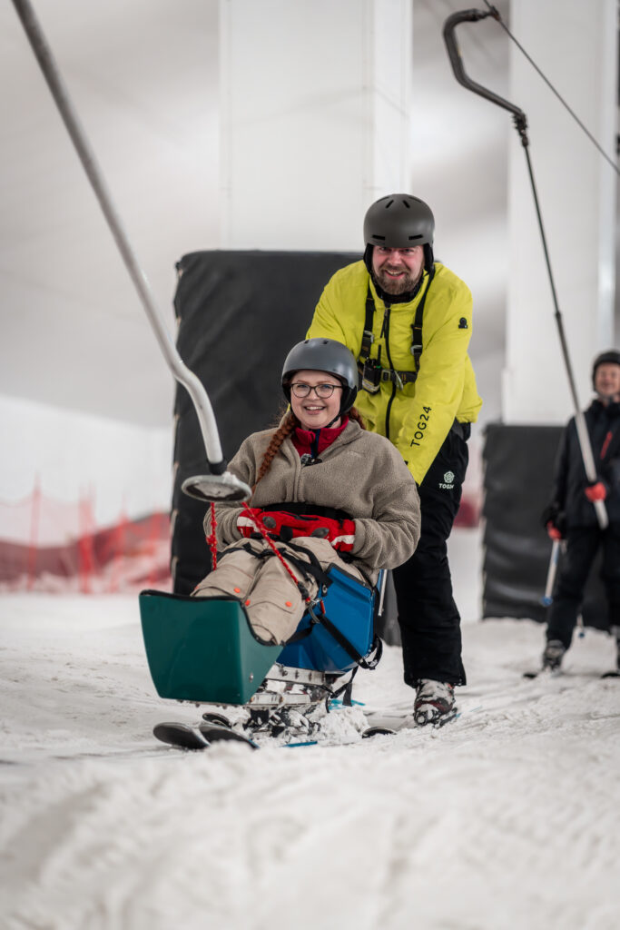 Jenni is sat in a sit ski, being pulled up a ski slope. Her instructor Mike behind her.