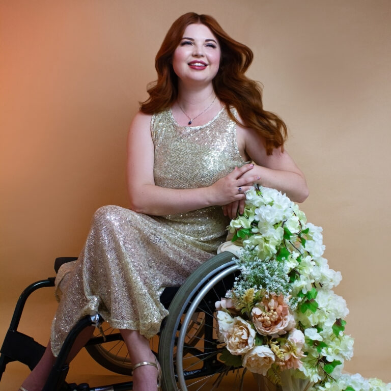 Jenni sat in her wheelchair, with flowers trailing off of it. She is wearing a gold sequin dress with heels.