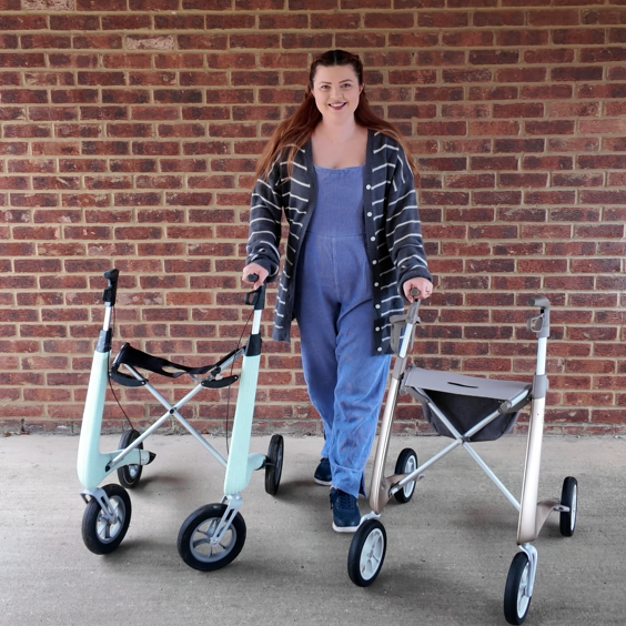 Jenni a white disabled woman with auburn hair is standing with one arm leaning on each rollator walker she is reviewing. Her right hand is on her pale mint green Nordic Pioneer whilst her left is on her champagne gold carbon ultralight. she is wearing a blue jumpsuit and grey cardigan.