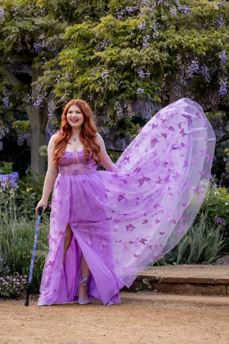 Jenni standing outdoors beneath cascading wisteria blossoms, smiling warmly while wearing a floral purple dress.