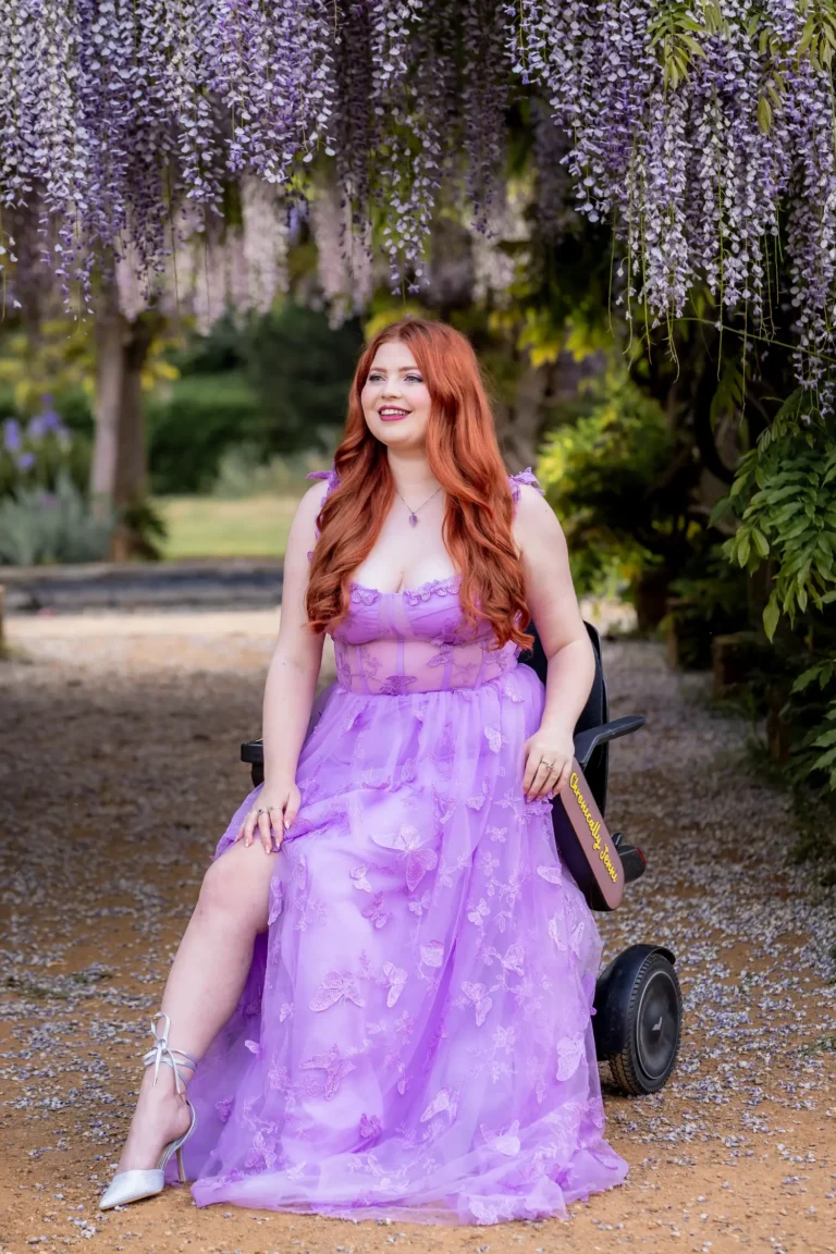 Jenni siting in her adapted wheelchair outdoors beneath cascading wisteria blossoms, smiling warmly while wearing a floral purple dress.