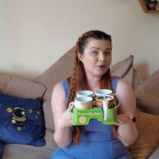 Jenni a white woman with auburn hair sat on her sofa wearing blue dunagrees holding a green gadgets which holds 4 mugs at once